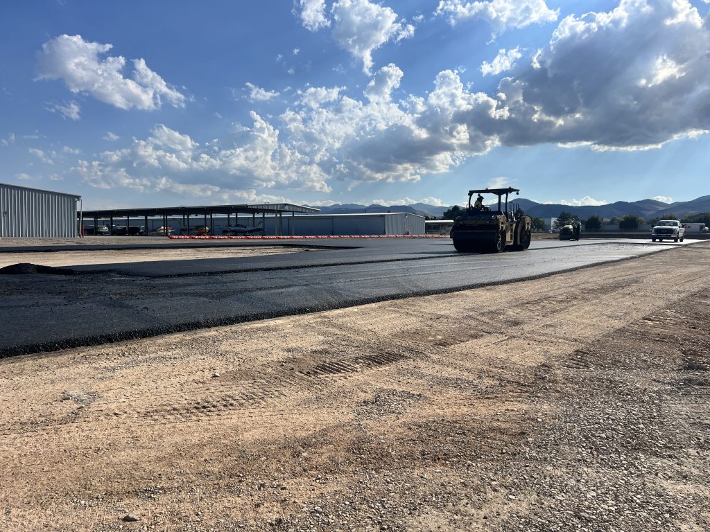 Paving equipment steamrolls hot asphalt near T-hangars.